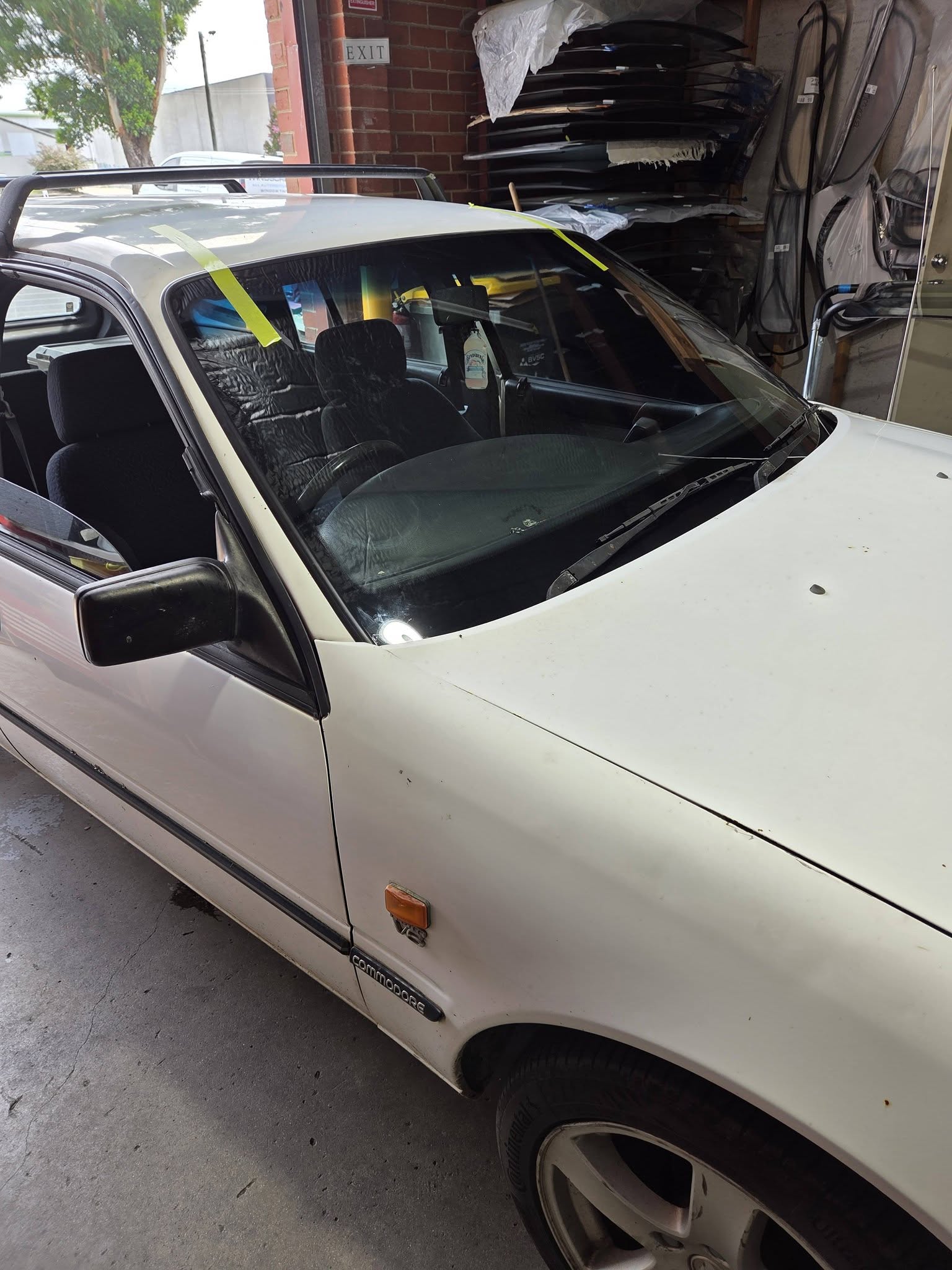 A white Holden Commodore inside the Blue Water Windscreens workshop with a freshly fitted windscreen held in place by tape, racks of glass stacked behind it