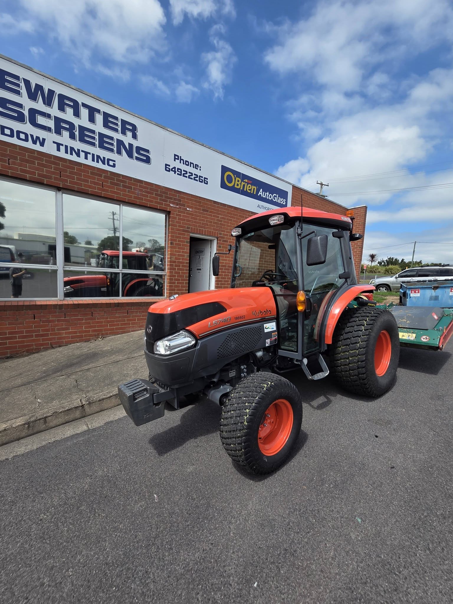 An orange Kubota L5740 tractor parked at the roller door of the Blue Water Windscreens workshop, with the window tinting sign above