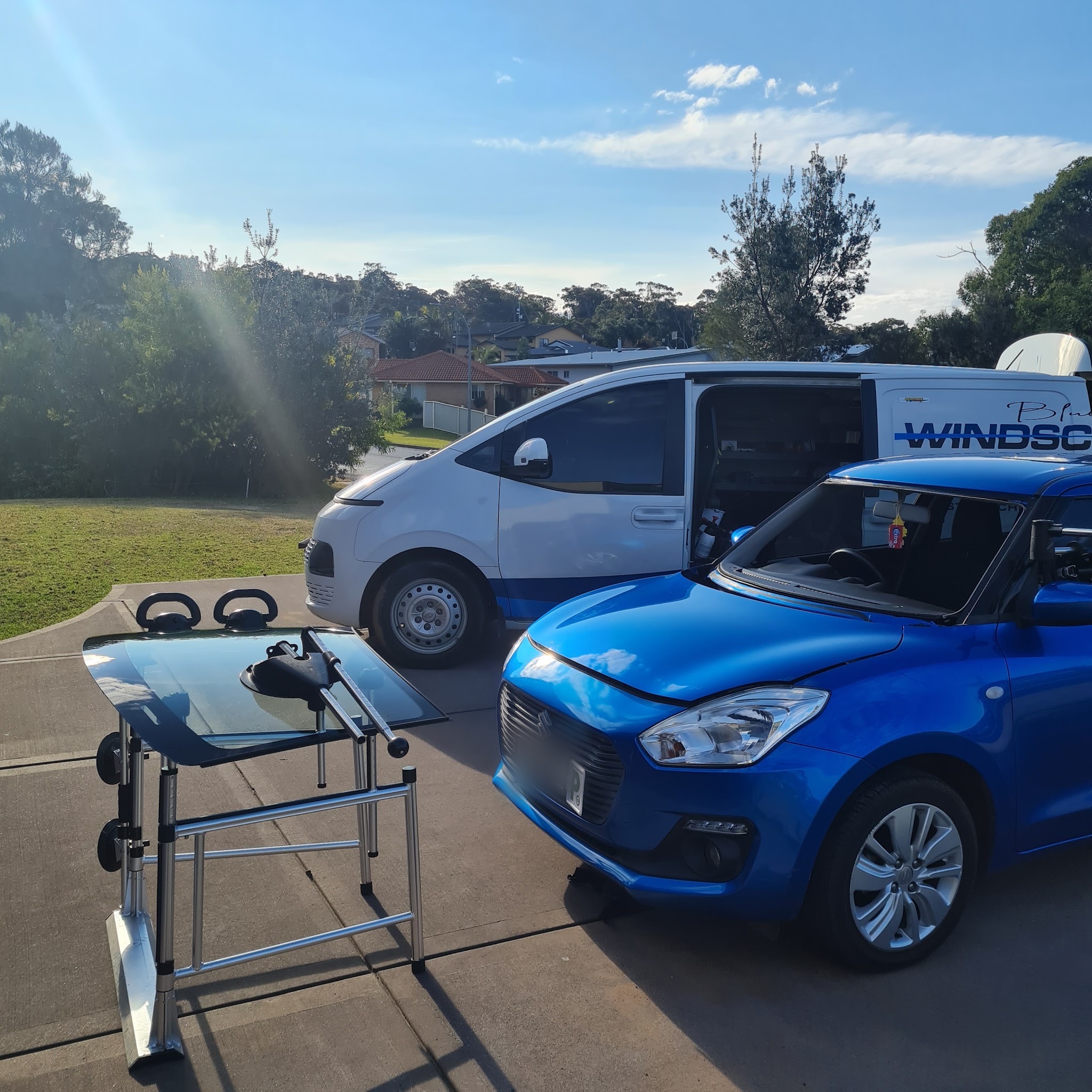 A blue Suzuki Swift with its windscreen removed, the replacement screen resting on a stand, and the Blue Water Windscreens van behind with its side door open